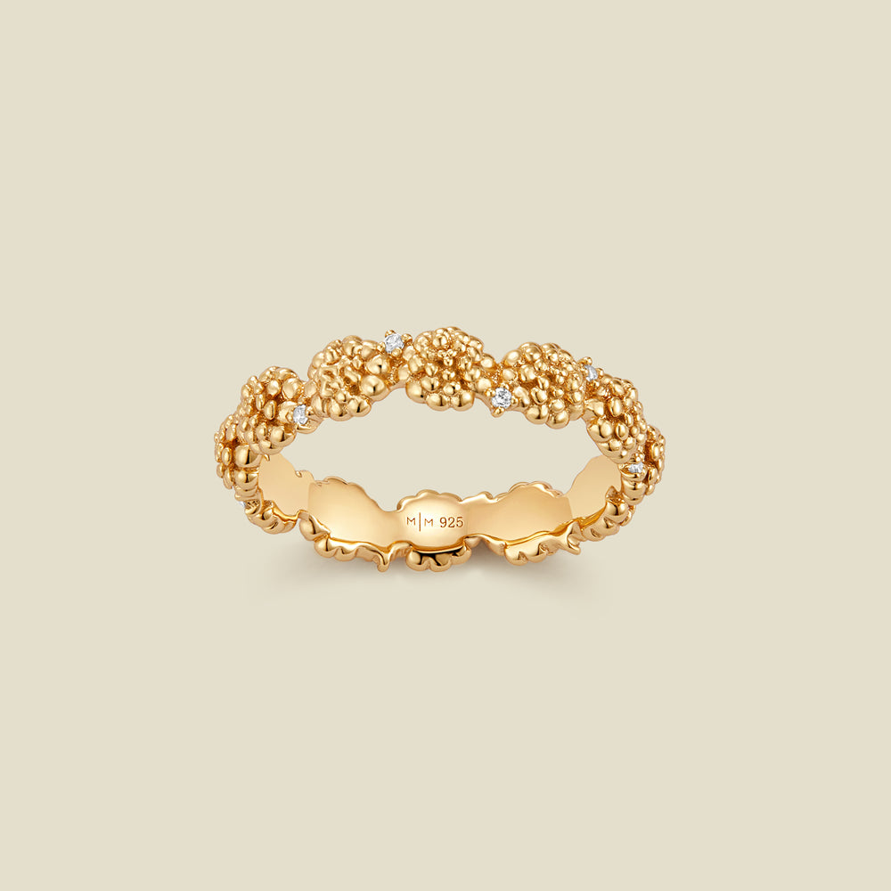 A gold floral-etched ring band with textured marigold flowers and small clear gemstones, shown on a plain warm beige background. 