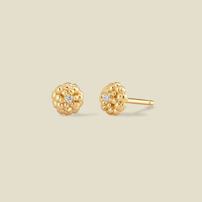 A pair of gold stud earrings with a textured, marigold flower-like cluster design and a small clear gemstone in the center of each, shown on a solid light beige background.