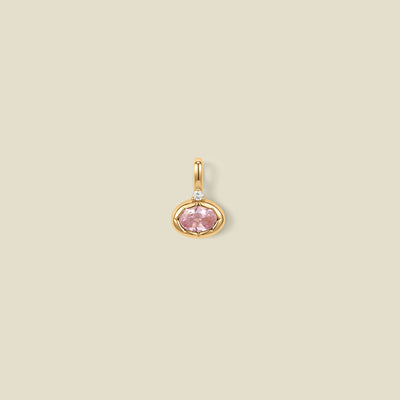A gold oval pendant charm with a pink bezel-set oval gemstone and a clear accent stone on top, set against a solid beige background.