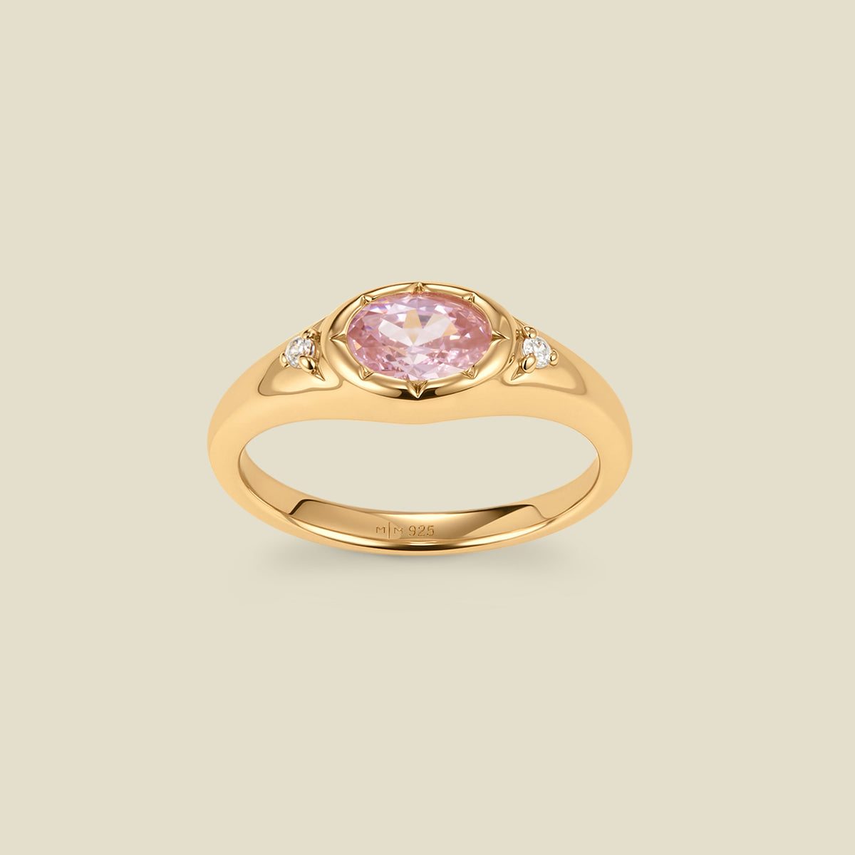 A gold signet ring with a pink center oval bezel-set gemstone, and two small clear accent stones on either side, set against a solid beige background.