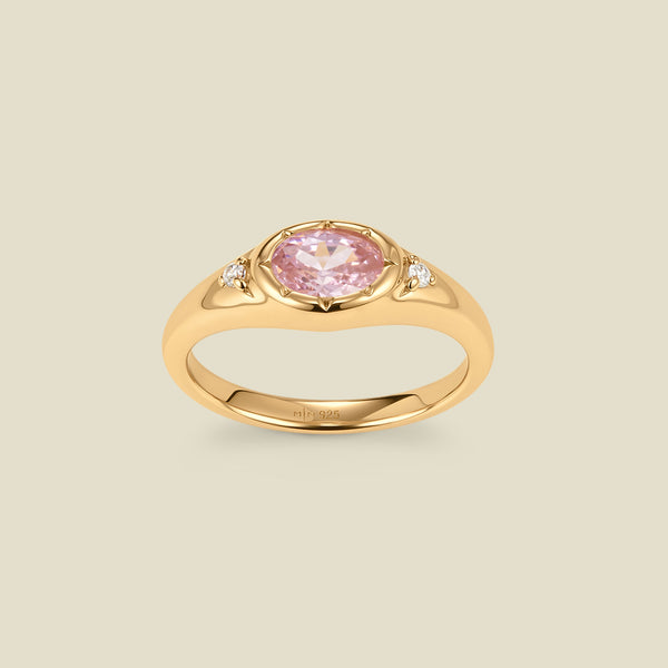 A gold signet ring with a pink center oval bezel-set gemstone, and two small clear accent stones on either side, set against a solid beige background.