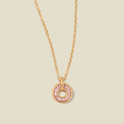 A gold chain necklace with a circular pendant featuring pink baguette-cut stones around a hollow center, displayed against a solid light beige background.