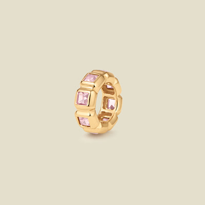 A small gold circular charm comprised of square, bezel-set pink gemstones side by side, set against a solid beige background.