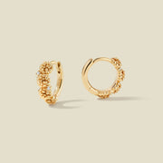 A pair of gold hoop earrings adorned with marigold flowers stacked along the hoop, with scattered small clear accent stones, set against a solid beige background.
