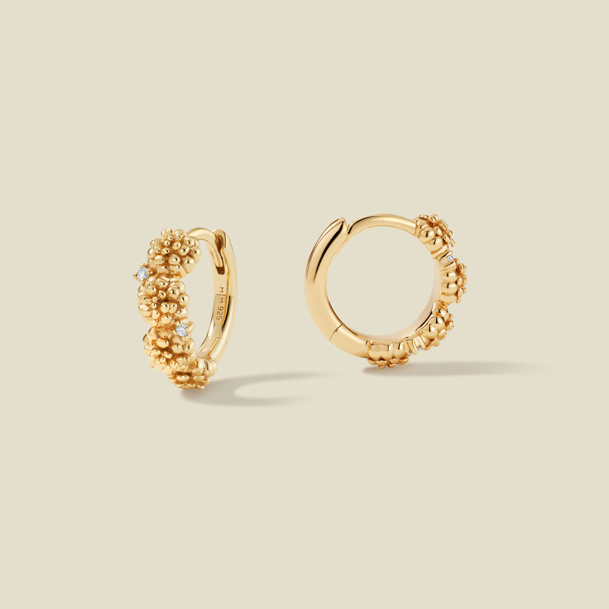 A pair of gold hoop earrings adorned with marigold flowers stacked along the hoop, with scattered small clear accent stones, set against a solid beige background.