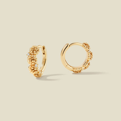 A pair of gold hoop earrings adorned with marigold flowers stacked along the hoop, with scattered small clear accent stones, set against a solid beige background.