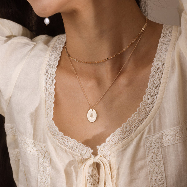 A close-up of a person wearing layered gold necklaces, including a delicate chain necklace and a longer chain with an oval pendant engraved with the a pine tree, styled with a cream lace blouse.