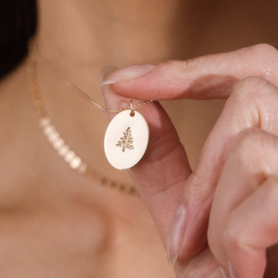 A close-up lifestyle shot of a person holding a delicate gold necklace with an oval pendant featuring an engraved pinr tree design. The background shows a soft-focus view of the person's skin and neck.