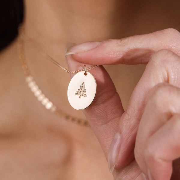 A close-up lifestyle shot of a person holding a delicate gold necklace with an oval pendant featuring an engraved pinr tree design. The background shows a soft-focus view of the person's skin and neck.