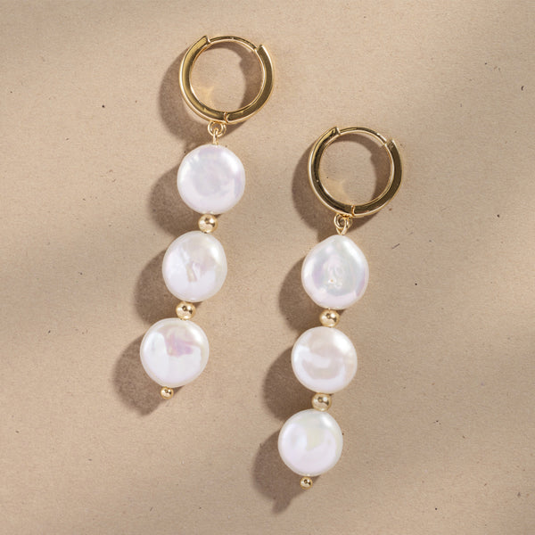 Gold hoop earrings with three dangling pearls against a plain beige background.
