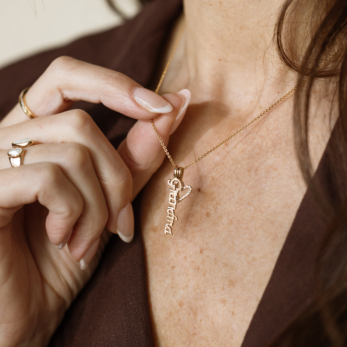 A close-up of a person wearing a delicate gold pendant chain necklace with a gold vertical charm spelling 'Grandma' and a gold heart outline charm covered in clear accent stones, paired with a brown top.