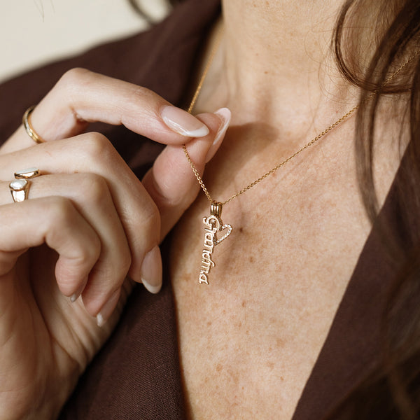 A close-up of a person wearing a delicate gold pendant chain necklace with a gold vertical charm spelling 'Grandma' and a gold heart outline charm covered in clear accent stones, paired with a brown top.