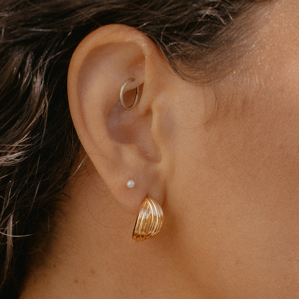 A close-up of an ear wearing a cross over textured gold hoop, a tiny pearl stud, and a small gold hoop.