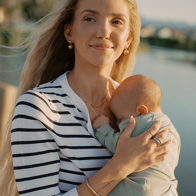 A woman holding a baby against her chest, wearing layered gold necklaces, multiple rings, a gold bangle bracelet and round bead dangle earrings, paired with a white and black striped cardigan, standing in front of a body of water.