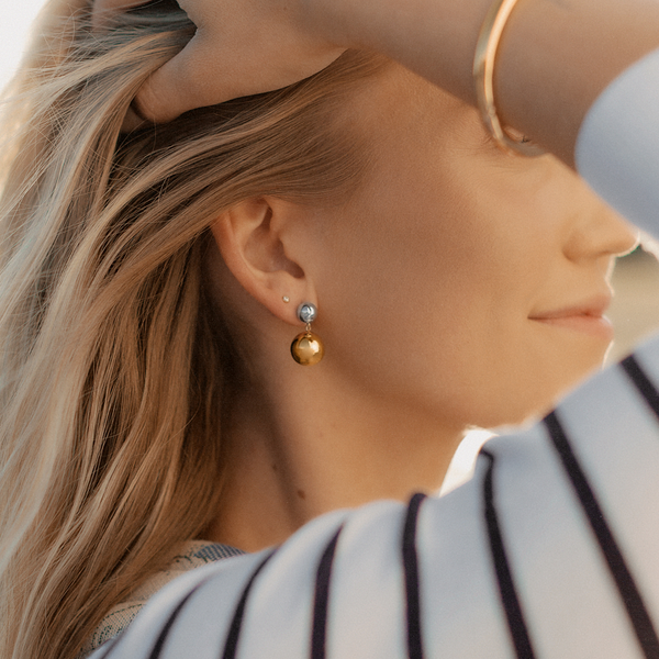 A close-up of a person's ear wearing a stud earring with a round silver ball and a larger round gold ball hanging from it.