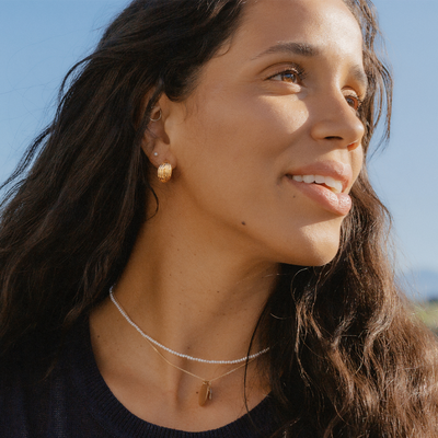 A woman wearing a black top with a pearl strand necklace layered with a gold chain necklace with multiple charms, and thick gold hoop earrings, set against a blue sky.