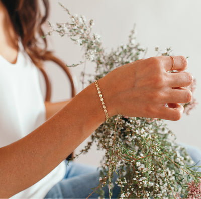 A close-up lifestyle shot of a woman's hand wearing a delicate gold chain bracelet, holding a bouquet of dried white flowers against a soft, light background.