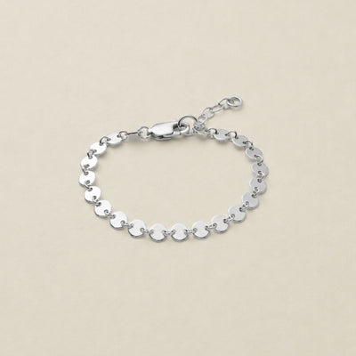 A silver chain bracelet made of flat, circular disc links, featuring a lobster clasp and extender chain, displayed on a light beige background.