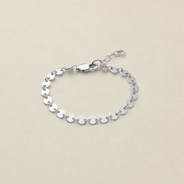 A silver chain bracelet made of flat, circular disc links, featuring a lobster clasp and extender chain, displayed on a light beige background.