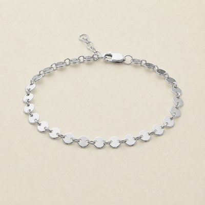 A silver chain bracelet made of flat, polished oval links, displayed on a light beige background.