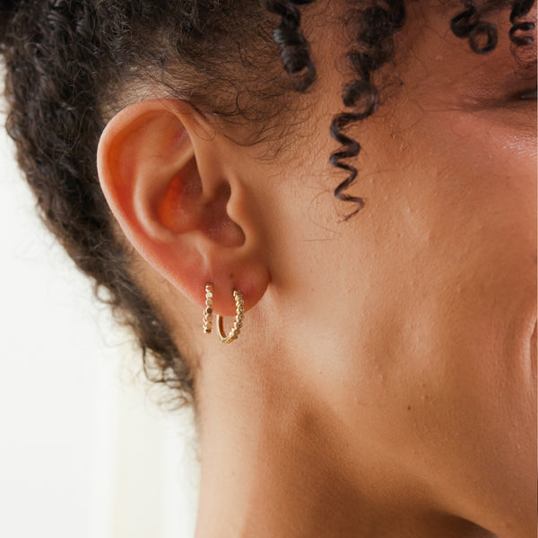 A close-up of a person's ear wearing a two small textured gold hoop earrings.