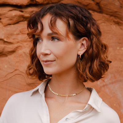 A woman with short curly red hair wearing a delicate gold chain necklace and a small hoop earring, posing in front of a warm, textured red rock background.