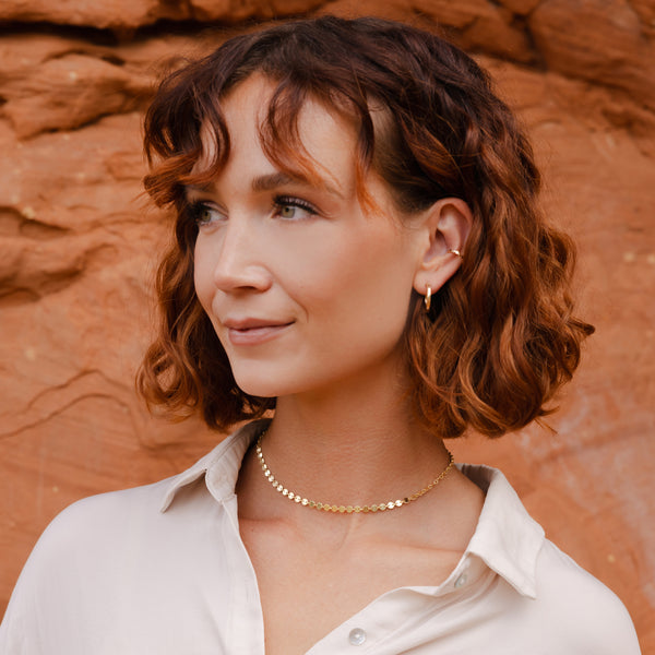 A woman with short curly red hair wearing a delicate gold chain necklace and a small hoop earring, posing in front of a warm, textured red rock background.