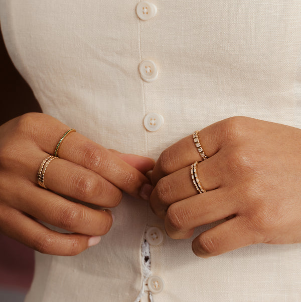 Close-up of a person's hands buttoning a cream-colored linen shirt, wearing delicate gold rings with small clear stones.