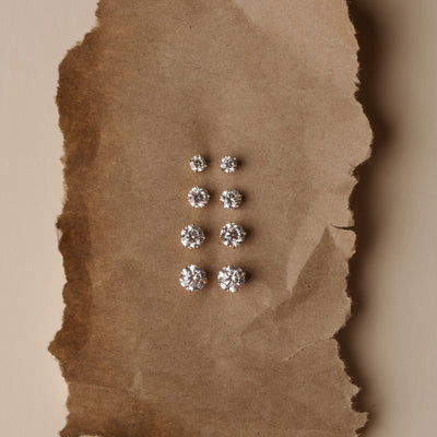 Six pairs of round brilliant-cut diamond stud earrings in silver settings, arranged vertically on a torn piece of brown kraft paper against a beige background.