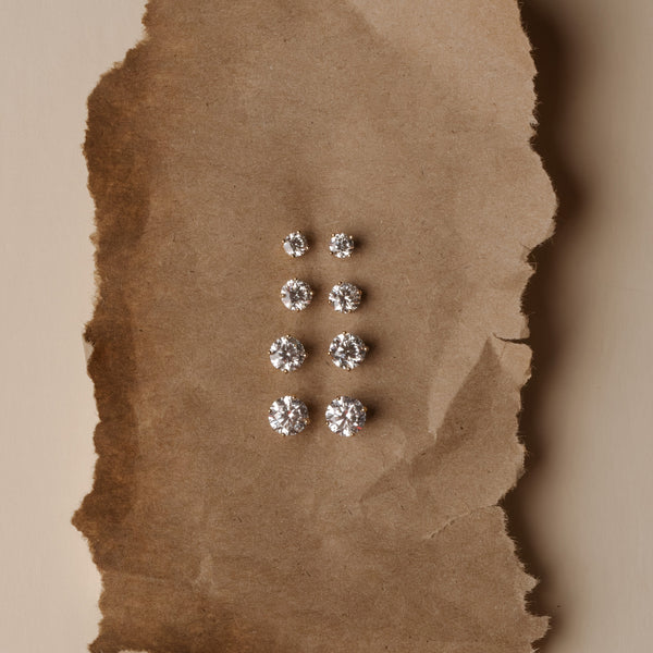 Six pairs of round brilliant-cut diamond stud earrings in silver settings, arranged vertically on a torn piece of brown kraft paper against a beige background.