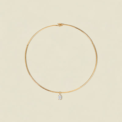 A thin gold wire hoop necklace with a small clear marquise gemstone pendant, displayed on a solid light beige background.