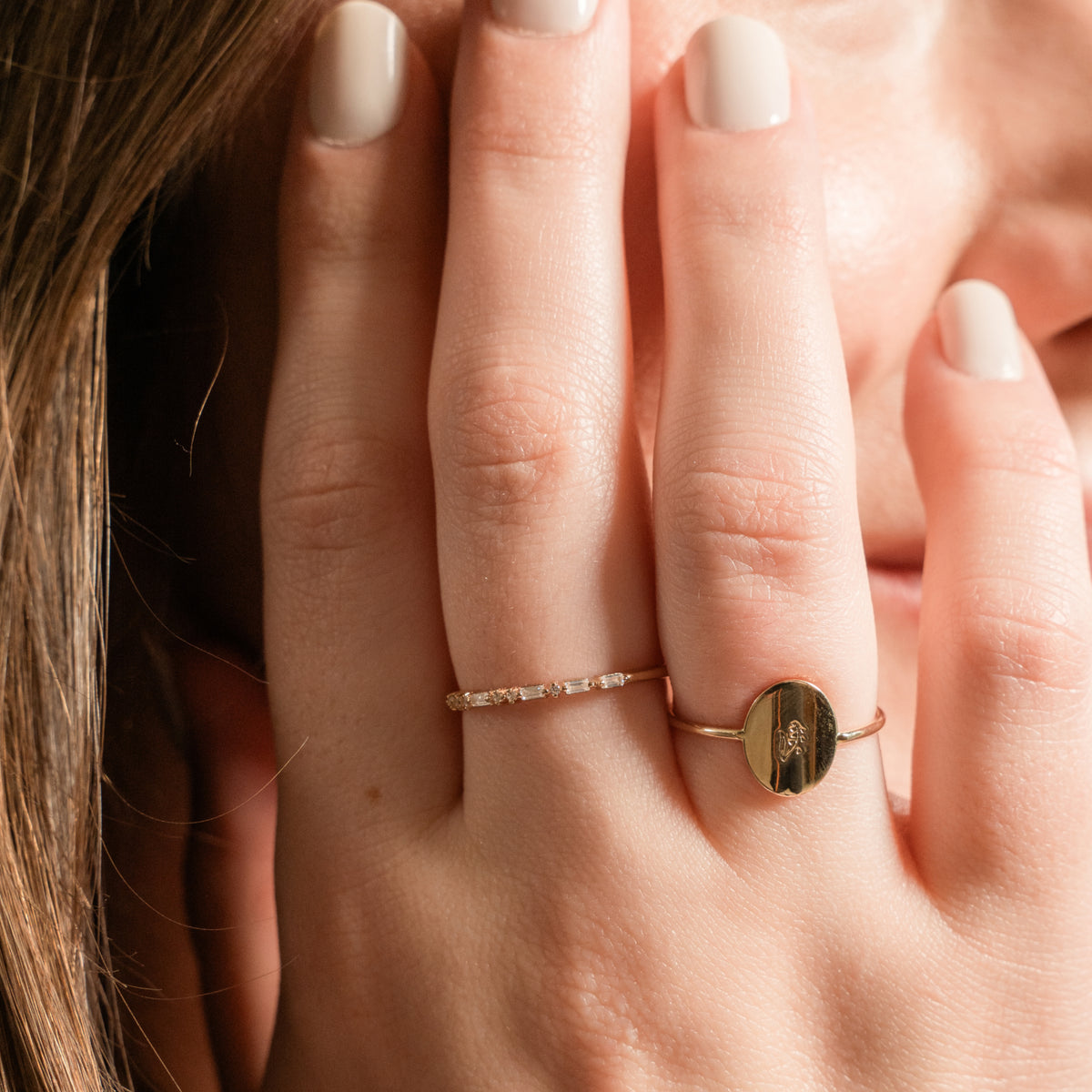 A close-up of a person's hand wearing two delicate gold rings: one with a row of small clear stones and the other featuring a polished oval gold disc.