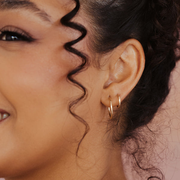 Close-up of a person's ear wearing two delicate gold hoop earrings, with dark curly hair and a soft pink background.
