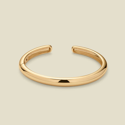 A polished gold-tone open cuff bangle bracelet, shown on a solid light beige background.