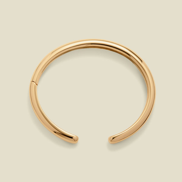 A polished gold open-end bangle bracelet, shown from a top angle, set against a solid beige background.