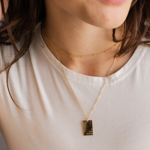 A close-up of a person wearing a delicate gold necklace with a rectangular dark gold pendant engraved with roman numerals.