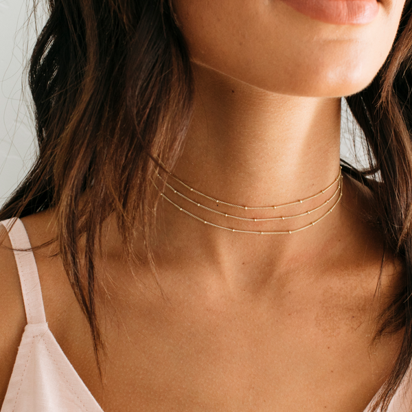 A close-up of a woman wearing three  delicate layered gold choker necklaces with small bead details.