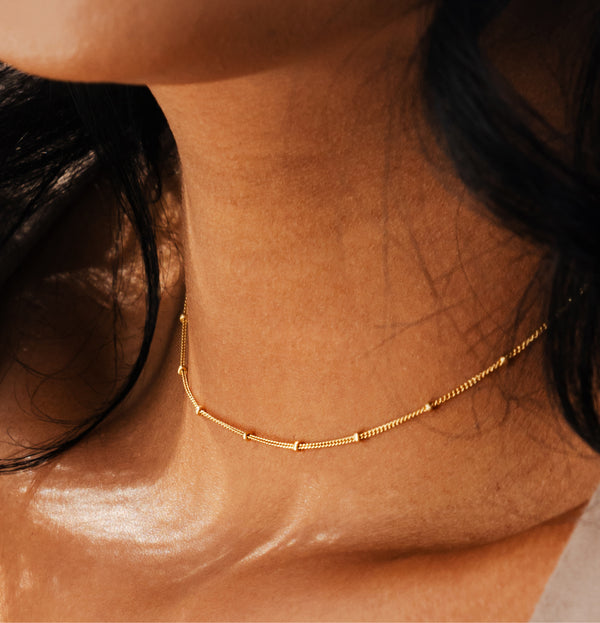 A close-up of a delicate gold chain necklace worn around a person's neck, featuring small spaced gold beads on a textured chain.