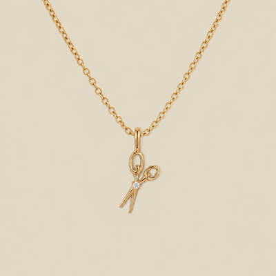 A delicate gold chain necklace featuring a small scissors pendant with a tiny clear gemstone at the pivot, displayed against a solid light beige background.