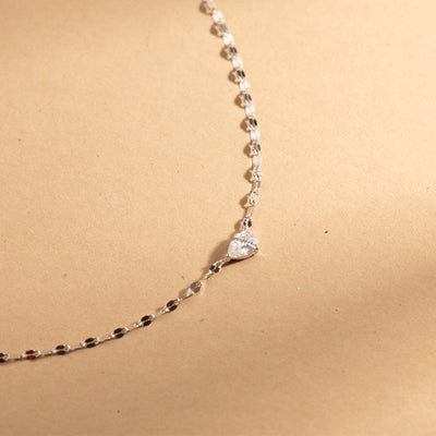 A dainty silver necklace with a large solitary tear drop shaped clear stone on it's side in the center, set against a warm textured beige background.
