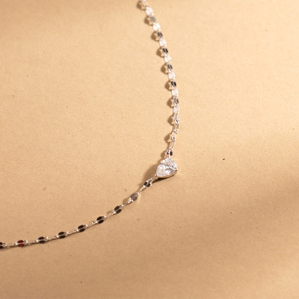 A dainty silver necklace with a large solitary tear drop shaped clear stone on it's side in the center, set against a warm textured beige background.