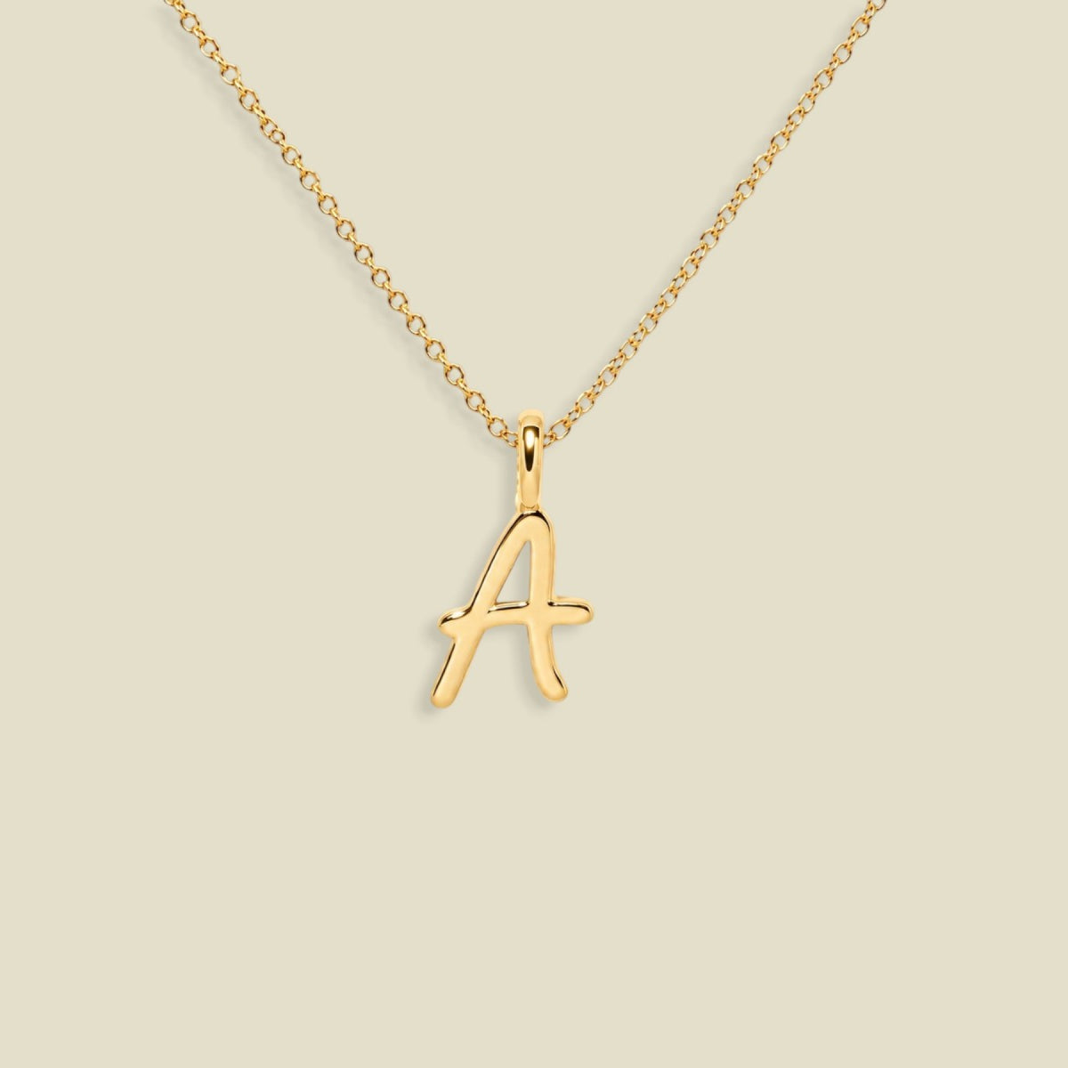 A gold initial necklace featuring a cursive, openwork letter 'A' pendant on a delicate gold chain, set against a solid light beige background.