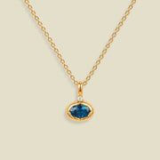 A gold chain necklace with a pendant featuring an oval dark blue-green gemstone in a gold bezel setting, accented by a small clear stone on the top, set against a solid light beige background.