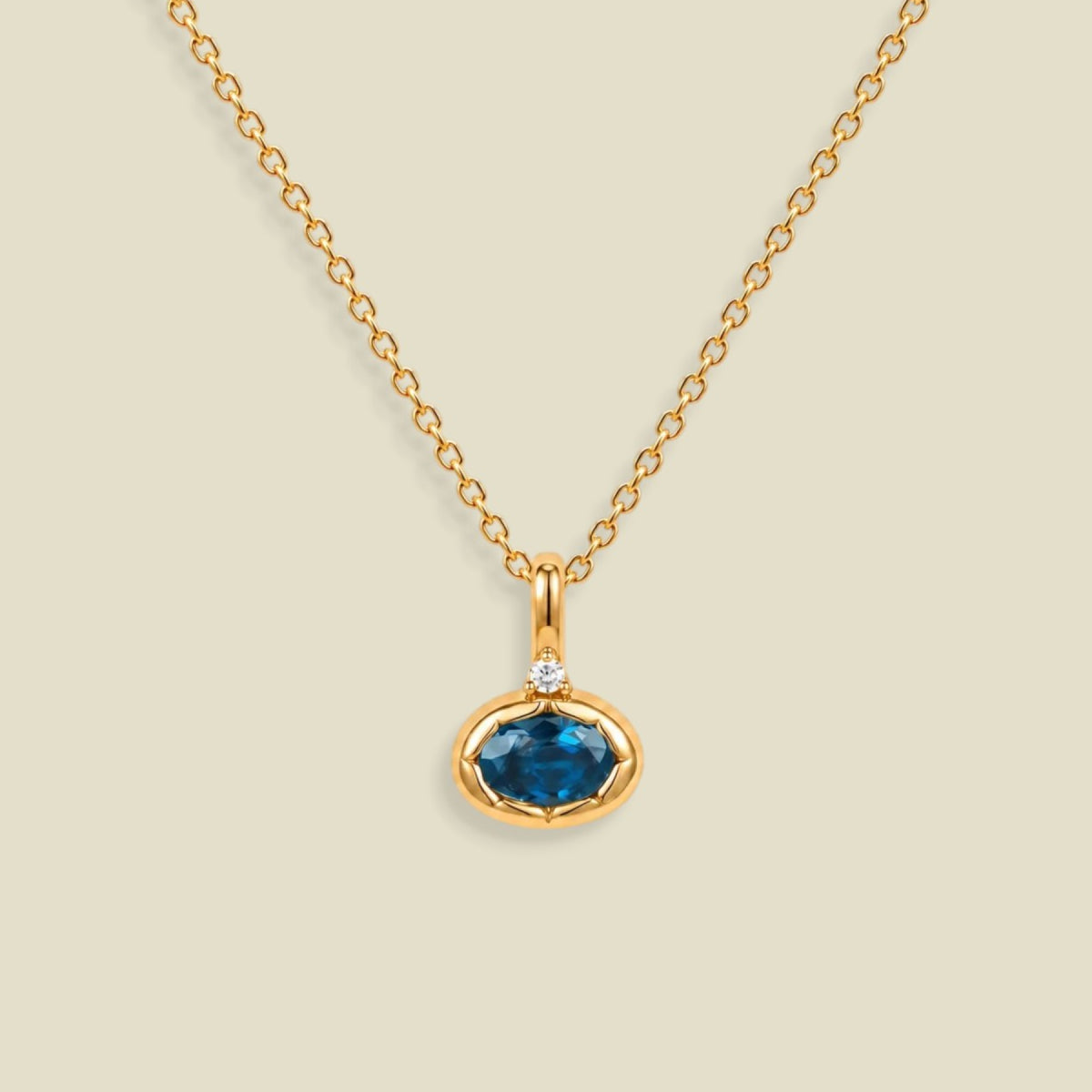 A gold chain necklace with a pendant featuring an oval dark blue-green gemstone in a gold bezel setting, accented by a small clear stone on the top, set against a solid light beige background.
