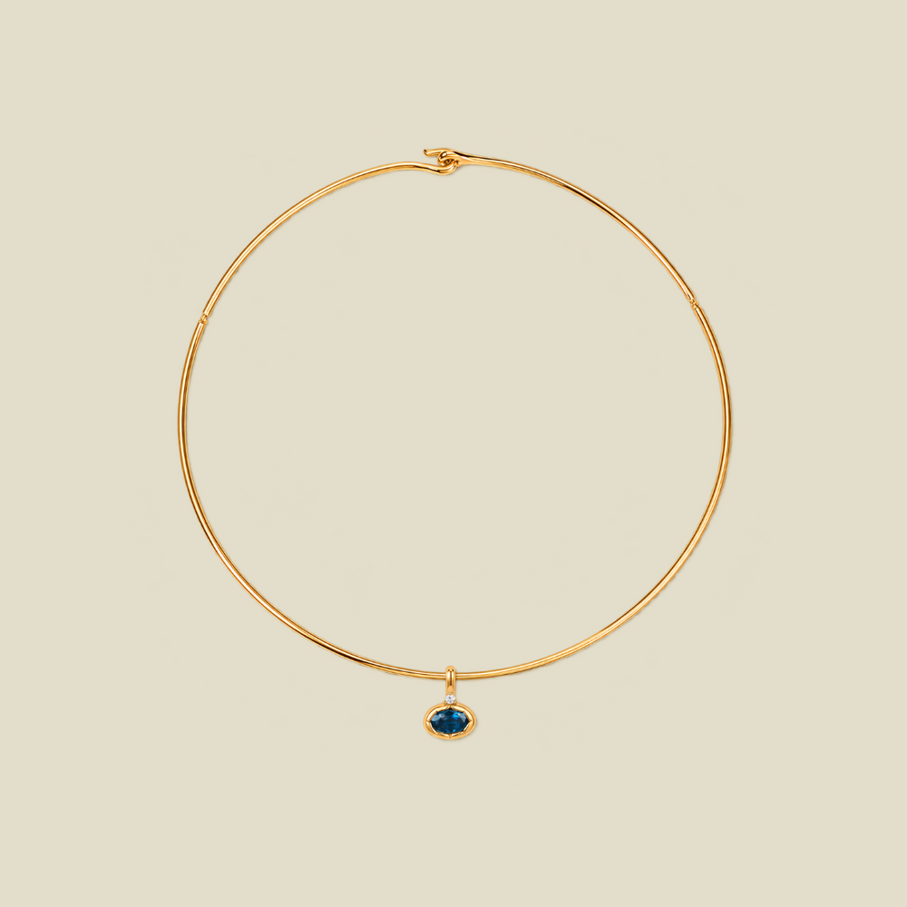 A thin gold wire hoop necklace with a small oval blue-green gemstone pendant, displayed on a solid light beige background.