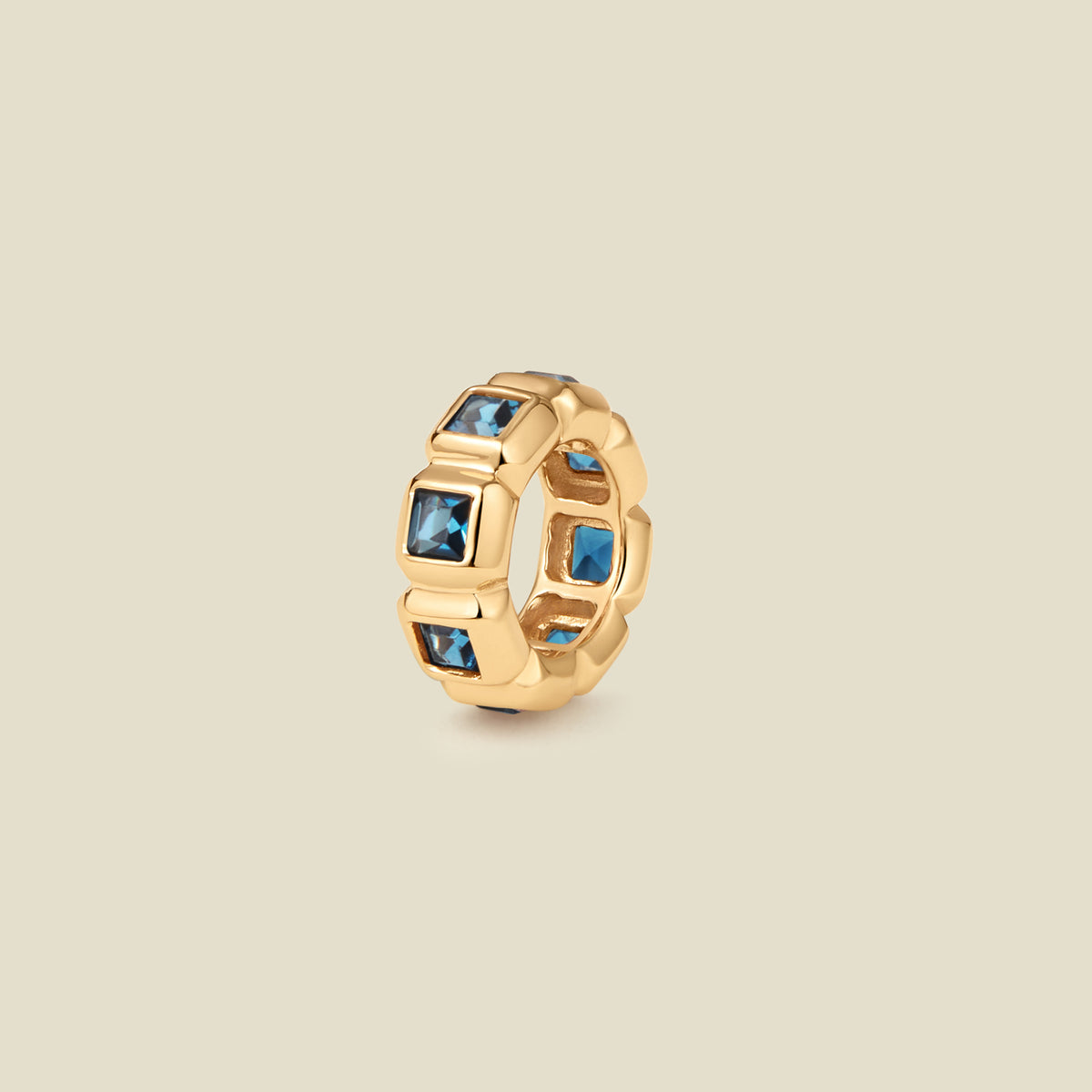 A gold ring charm with multiple bezel set square-cut blue gemstones set side by side to create the band, shown on a solid beige background.