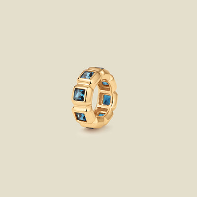 A gold ring charm with multiple bezel set square-cut blue gemstones set side by side to create the band, shown on a solid beige background.