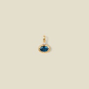 A gold oval pendant charm with a blue-green bezel-set oval gemstone and a clear accent stone on top, set against a solid beige background.