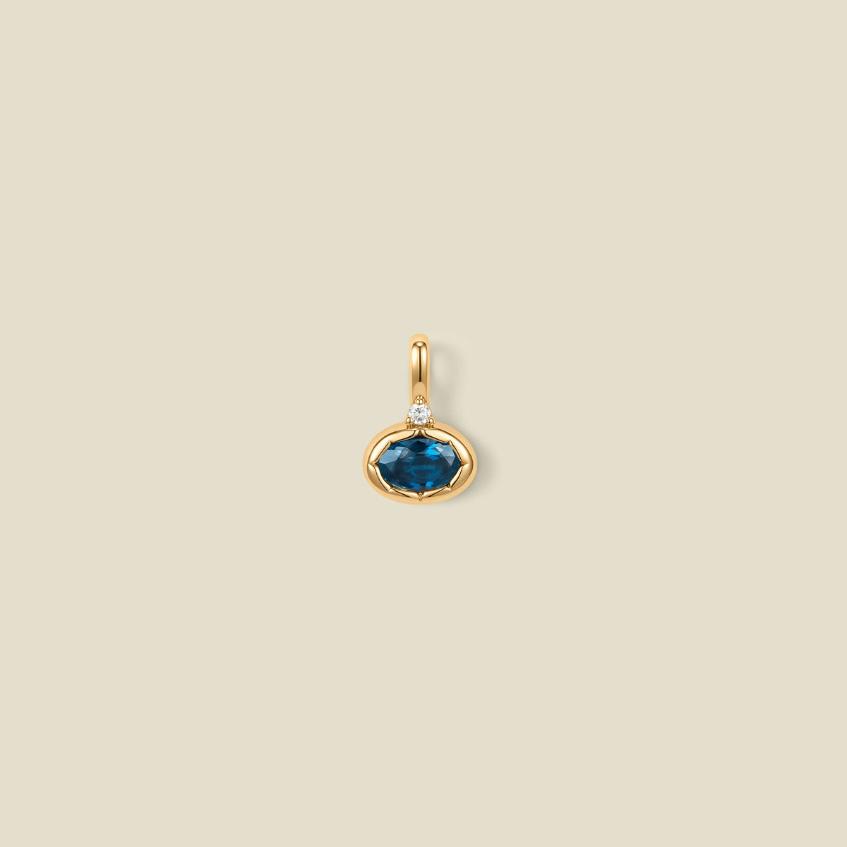 A gold oval pendant charm with a blue-green bezel-set oval gemstone and a clear accent stone on top, set against a solid beige background.