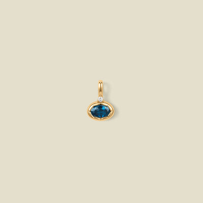 A gold oval pendant charm with a blue-green bezel-set oval gemstone and a clear accent stone on top, set against a solid beige background.